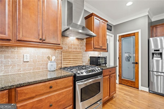 kitchen featuring wall chimney exhaust hood, stainless steel appliances, dark stone counters, light hardwood / wood-style floors, and ornamental molding