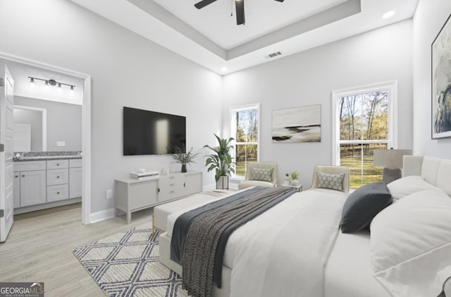 bedroom featuring ceiling fan, a tray ceiling, ensuite bath, and light hardwood / wood-style flooring