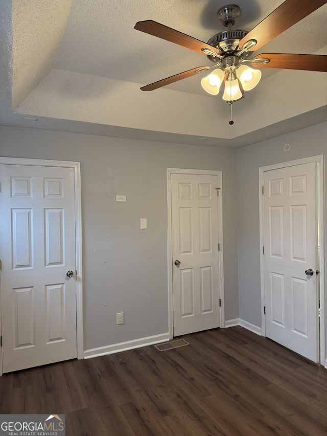 unfurnished bedroom featuring a textured ceiling, ceiling fan, dark wood-type flooring, and a tray ceiling