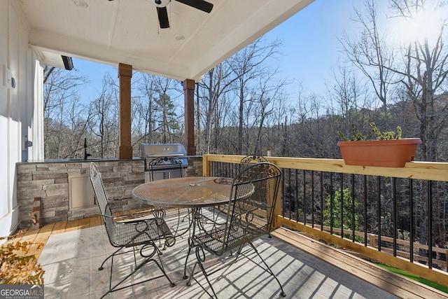 wooden terrace with outdoor dining space, a ceiling fan, a forest view, a grill, and exterior kitchen