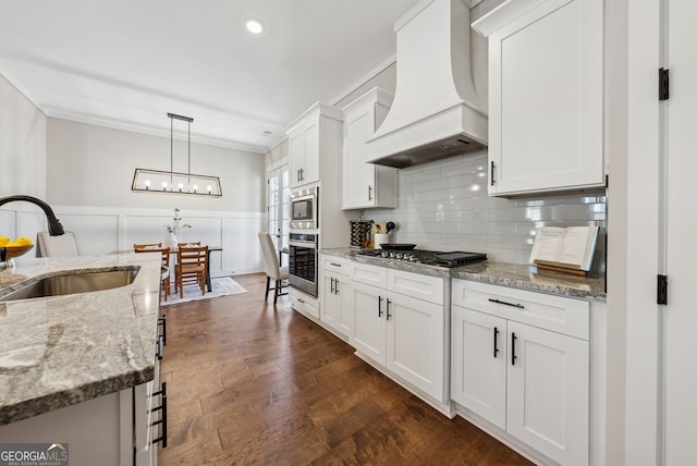 kitchen with premium range hood, wainscoting, appliances with stainless steel finishes, white cabinetry, and a sink