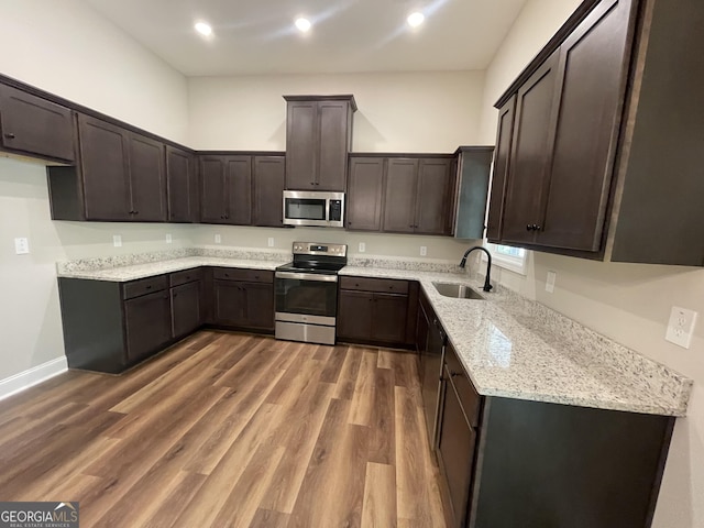 kitchen with dark brown cabinetry, appliances with stainless steel finishes, wood finished floors, a sink, and light stone counters