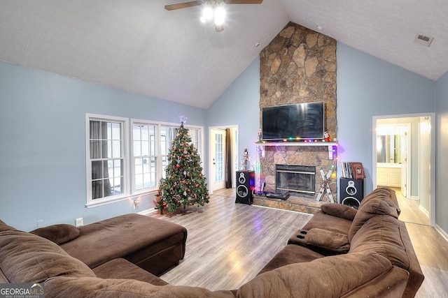 living room with high vaulted ceiling, a stone fireplace, ceiling fan, light wood-type flooring, and a textured ceiling