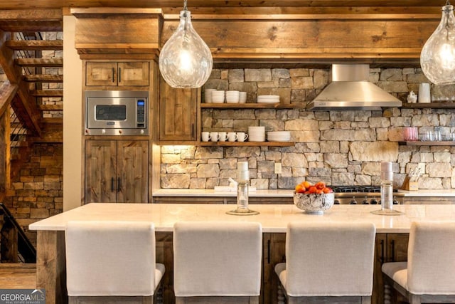 kitchen featuring a kitchen bar, wall chimney exhaust hood, decorative light fixtures, a kitchen island, and stainless steel microwave