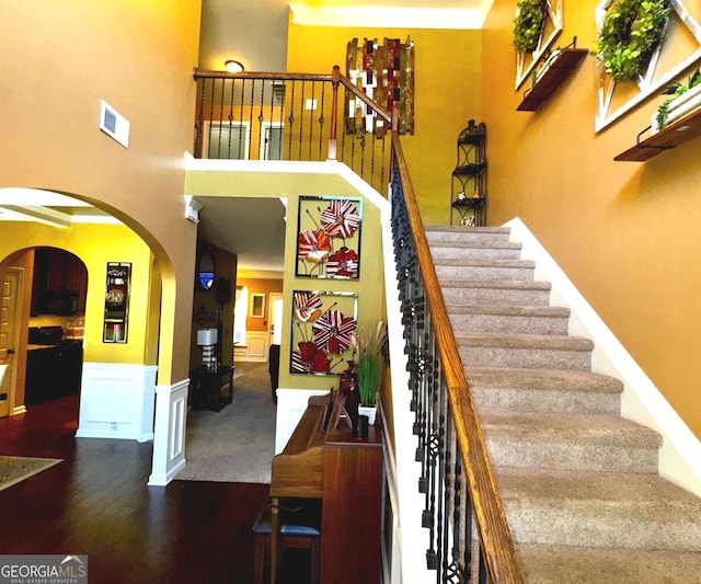 stairway featuring wood finished floors, arched walkways, a towering ceiling, wainscoting, and a decorative wall