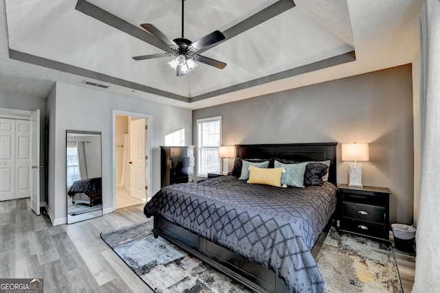 bedroom featuring a raised ceiling, ceiling fan, ensuite bathroom, and light wood-type flooring