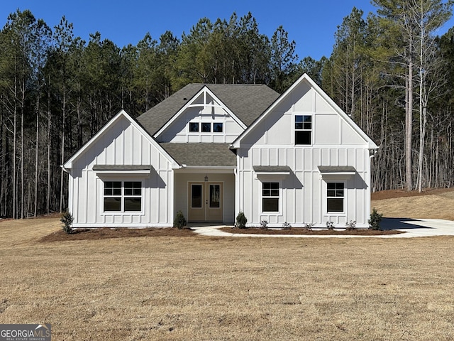 modern farmhouse style home with roof with shingles, board and batten siding, a front yard, and a forest view