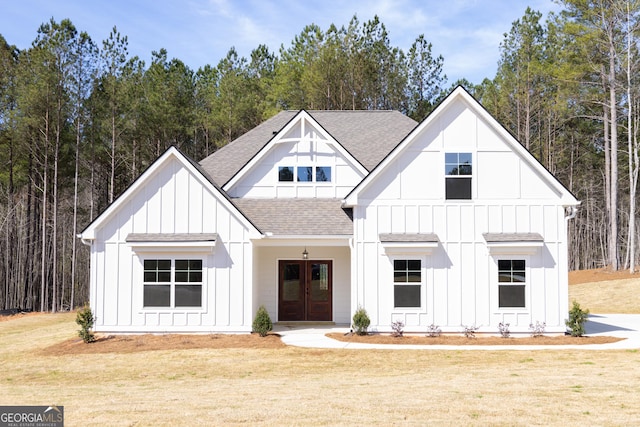 modern inspired farmhouse with french doors, board and batten siding, a forest view, a front yard, and a shingled roof