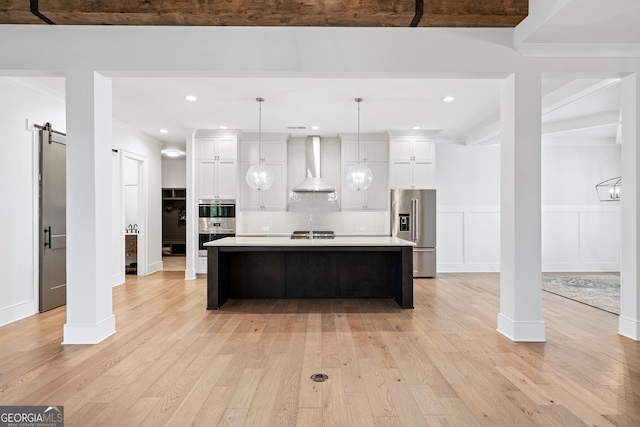 kitchen with stainless steel refrigerator with ice dispenser, wall chimney exhaust hood, pendant lighting, a barn door, and an island with sink