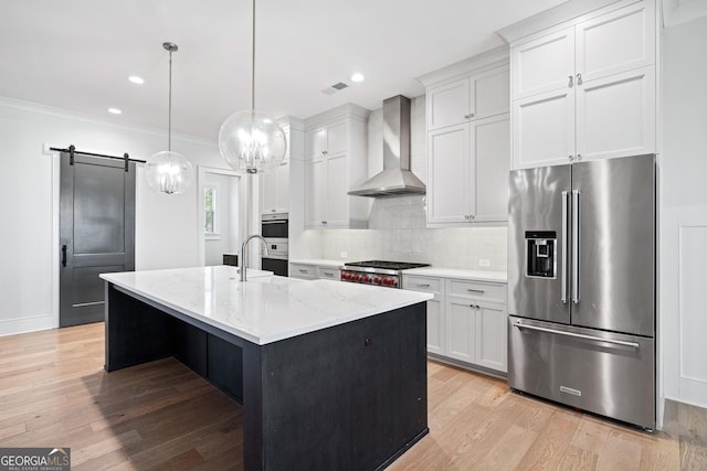 kitchen with white cabinetry, wall chimney range hood, a barn door, a center island with sink, and appliances with stainless steel finishes