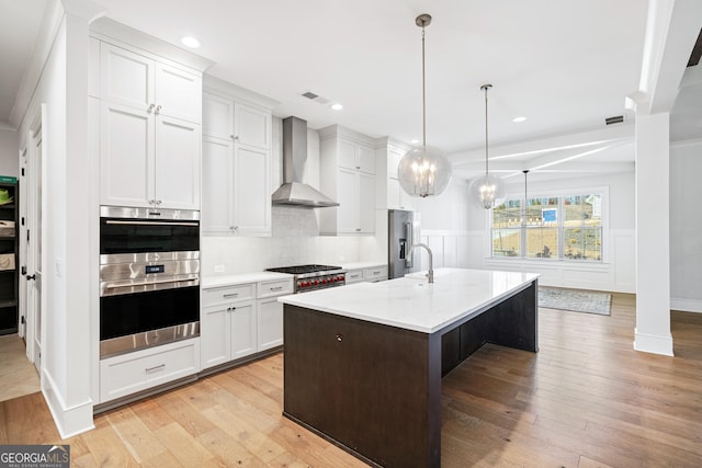 kitchen featuring a kitchen island with sink, white cabinets, wall chimney range hood, decorative light fixtures, and stainless steel appliances