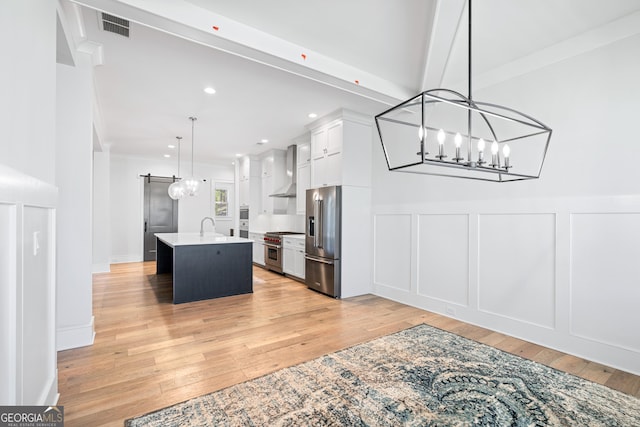 kitchen featuring premium appliances, wall chimney range hood, a center island with sink, white cabinetry, and hanging light fixtures