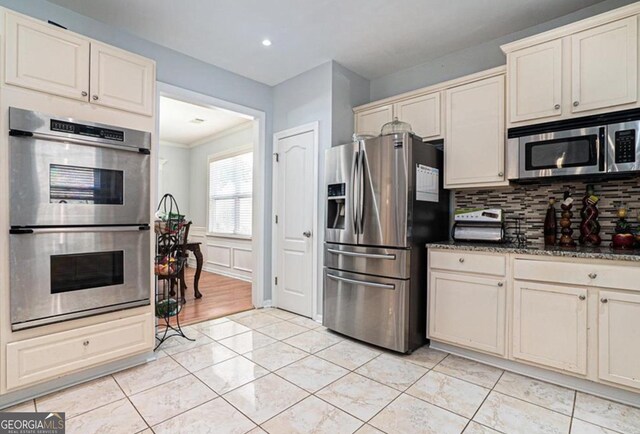 kitchen with stainless steel appliances, dark stone countertops, cream cabinets, decorative backsplash, and ornamental molding