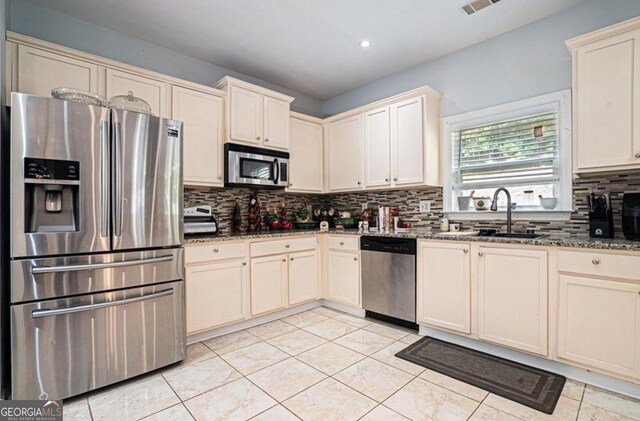 kitchen featuring sink, backsplash, dark stone countertops, cream cabinets, and appliances with stainless steel finishes