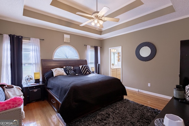 bedroom with light wood-type flooring, a raised ceiling, ceiling fan, crown molding, and connected bathroom