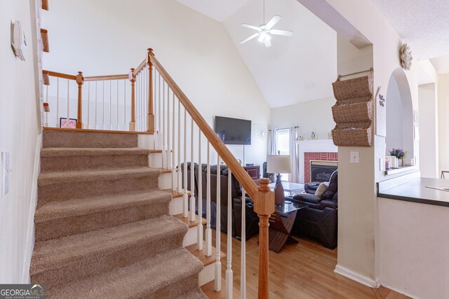 stairs featuring wood-type flooring, high vaulted ceiling, ceiling fan, and a brick fireplace