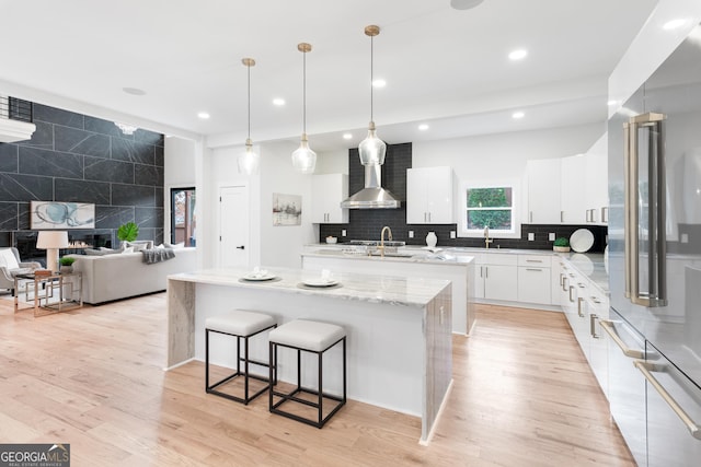 kitchen featuring light wood-type flooring, wall chimney range hood, decorative light fixtures, white cabinets, and a center island