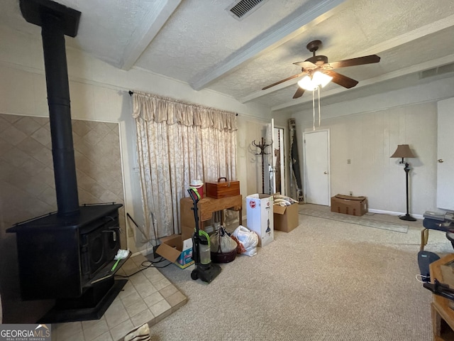 interior space with a textured ceiling, ceiling fan, beam ceiling, carpet floors, and a wood stove