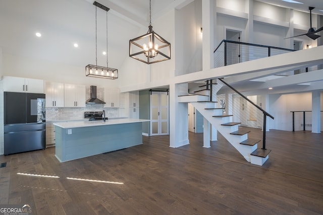 kitchen featuring black fridge, wall chimney range hood, hanging light fixtures, a towering ceiling, and white cabinetry