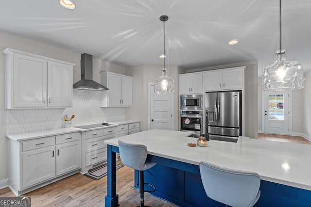 kitchen featuring appliances with stainless steel finishes, light wood-type flooring, wall chimney range hood, white cabinetry, and hanging light fixtures