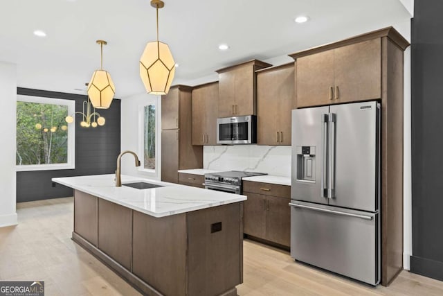 kitchen featuring light hardwood / wood-style floors, pendant lighting, appliances with stainless steel finishes, an island with sink, and light stone counters