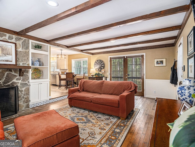 living area featuring beam ceiling, a wainscoted wall, french doors, light wood-style floors, and a stone fireplace