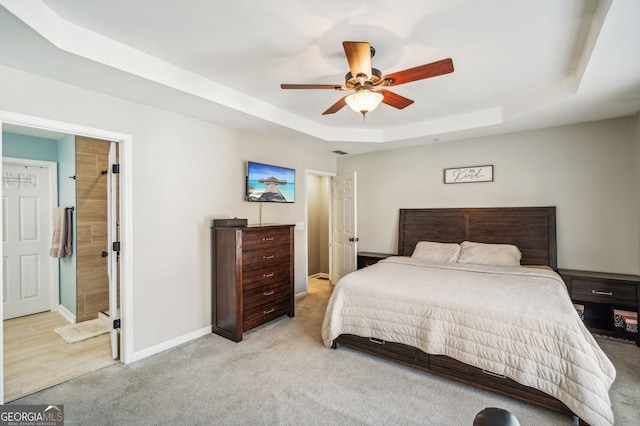 bedroom with ceiling fan, light colored carpet, baseboards, a raised ceiling, and ensuite bath