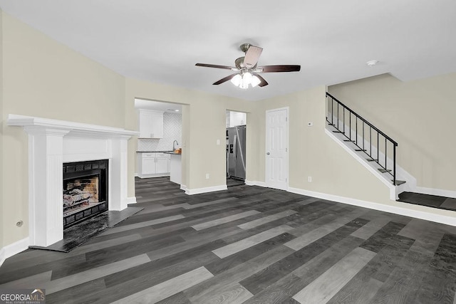 unfurnished living room with a fireplace, a ceiling fan, stairs, baseboards, and dark wood-style floors