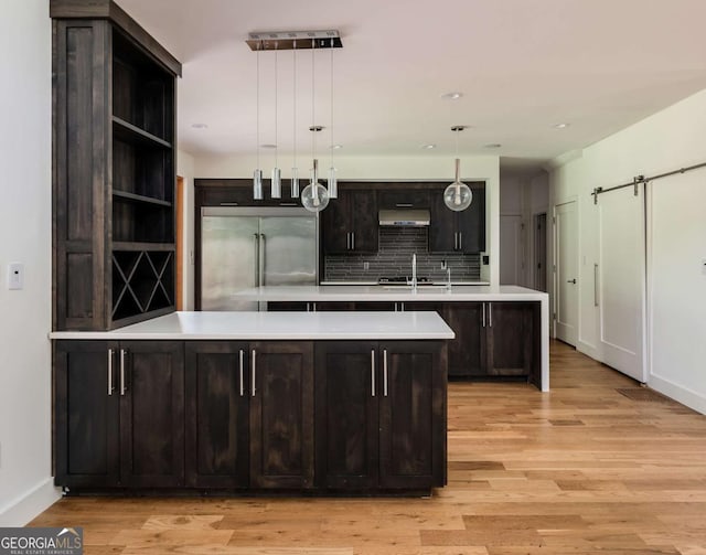 kitchen with hanging light fixtures, backsplash, built in refrigerator, dark brown cabinets, and light wood-type flooring