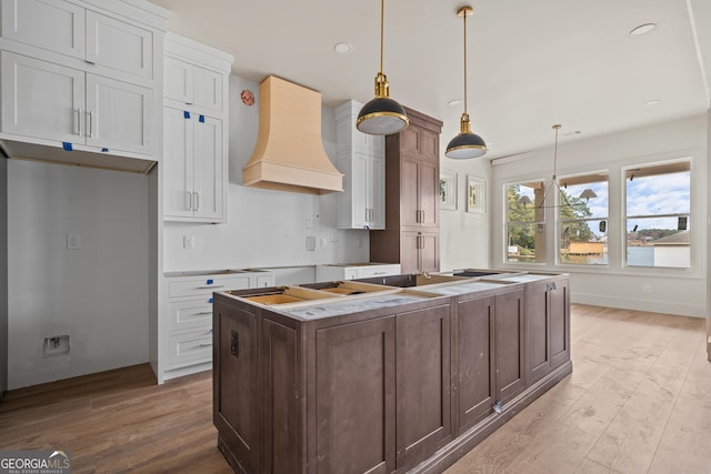 kitchen with light wood-type flooring, custom exhaust hood, a center island with sink, white cabinetry, and hanging light fixtures