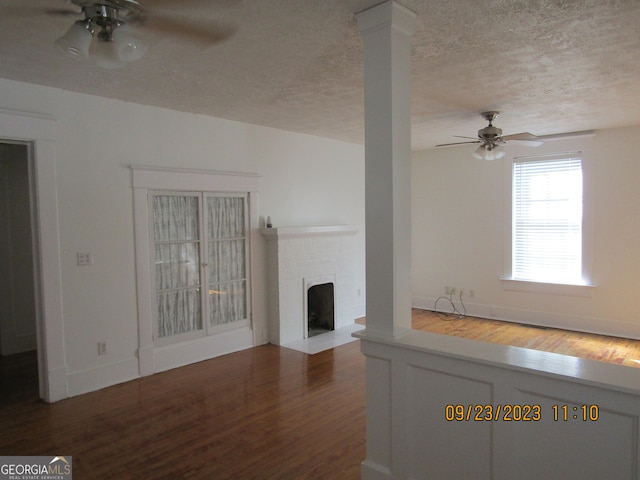 unfurnished living room featuring a brick fireplace, ceiling fan, ornate columns, a textured ceiling, and dark hardwood / wood-style flooring