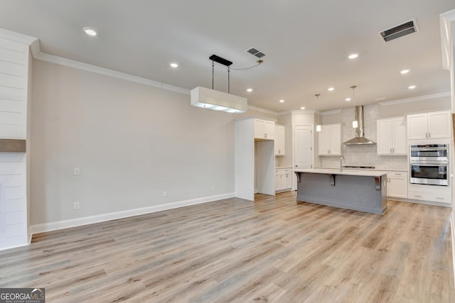 kitchen with wall chimney exhaust hood, double oven, a kitchen island with sink, light hardwood / wood-style flooring, and hanging light fixtures