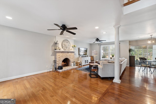 living room with ceiling fan with notable chandelier, wood-type flooring, a fireplace, and decorative columns