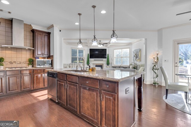 kitchen with a center island with sink, wall chimney range hood, sink, decorative light fixtures, and stainless steel appliances