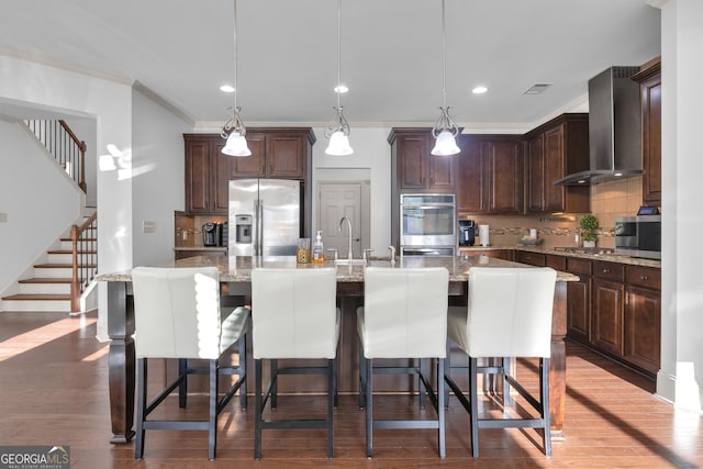 kitchen with backsplash, wall chimney range hood, an island with sink, and appliances with stainless steel finishes