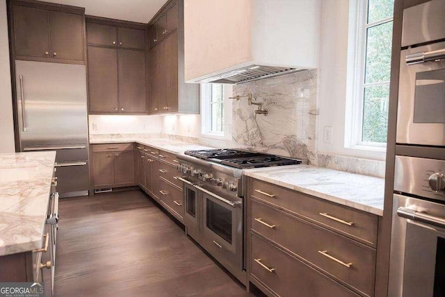 kitchen featuring dark wood-type flooring, wall chimney range hood, decorative backsplash, premium appliances, and light stone counters