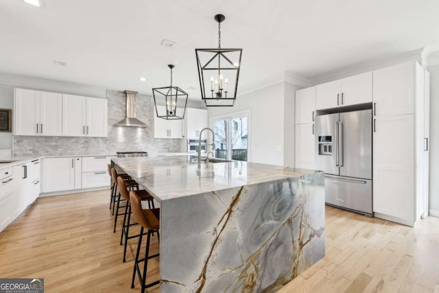 kitchen featuring high end refrigerator, wall chimney exhaust hood, sink, a center island with sink, and light hardwood / wood-style flooring