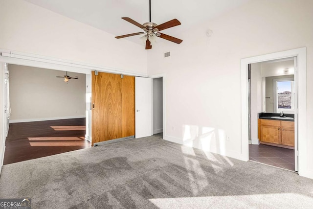 unfurnished bedroom featuring dark carpet, ensuite bathroom, sink, ceiling fan, and a towering ceiling