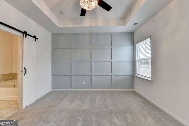 unfurnished bedroom with light carpet, a tray ceiling, ceiling fan, a barn door, and connected bathroom