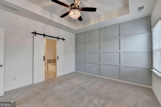 unfurnished bedroom with ensuite bath, ceiling fan, a raised ceiling, a barn door, and light carpet