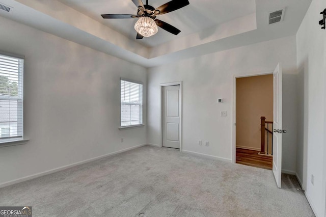 unfurnished bedroom with light colored carpet, multiple windows, a tray ceiling, and ceiling fan