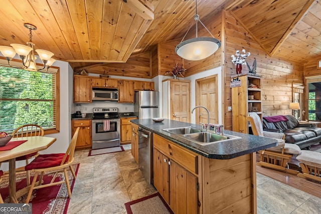 kitchen with sink, stainless steel appliances, wood walls, decorative light fixtures, and a center island with sink