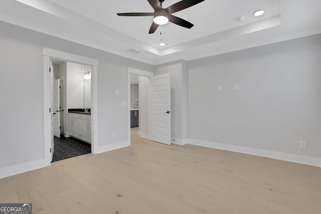 unfurnished bedroom with light hardwood / wood-style flooring, ensuite bath, ceiling fan, and a tray ceiling