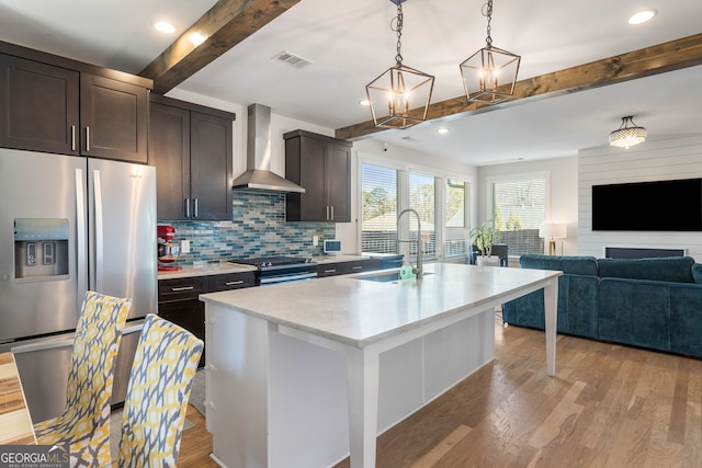 kitchen with appliances with stainless steel finishes, wall chimney exhaust hood, a kitchen island with sink, beamed ceiling, and hanging light fixtures