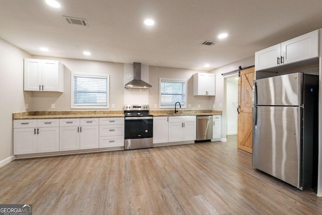 kitchen featuring appliances with stainless steel finishes, wall chimney range hood, a barn door, light hardwood / wood-style flooring, and white cabinets