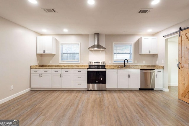 kitchen with white cabinets, appliances with stainless steel finishes, a barn door, and wall chimney range hood