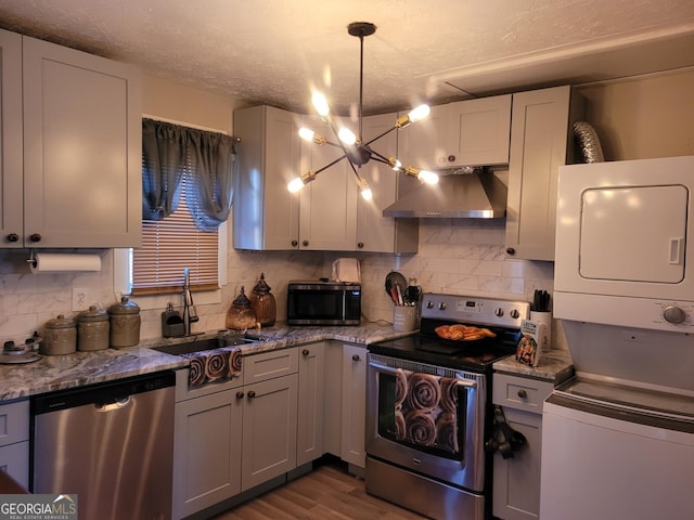 kitchen with wall chimney range hood, sink, light wood-type flooring, appliances with stainless steel finishes, and stacked washer / dryer