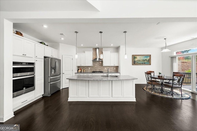 kitchen featuring appliances with stainless steel finishes, wall chimney exhaust hood, decorative light fixtures, white cabinetry, and an island with sink