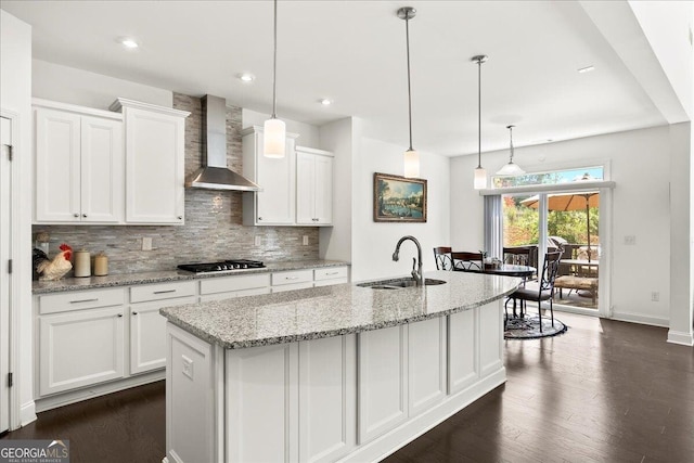 kitchen with a kitchen island with sink, white cabinets, hanging light fixtures, and wall chimney range hood