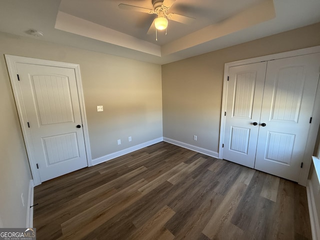 unfurnished bedroom with a tray ceiling, dark wood-style flooring, a closet, a ceiling fan, and baseboards